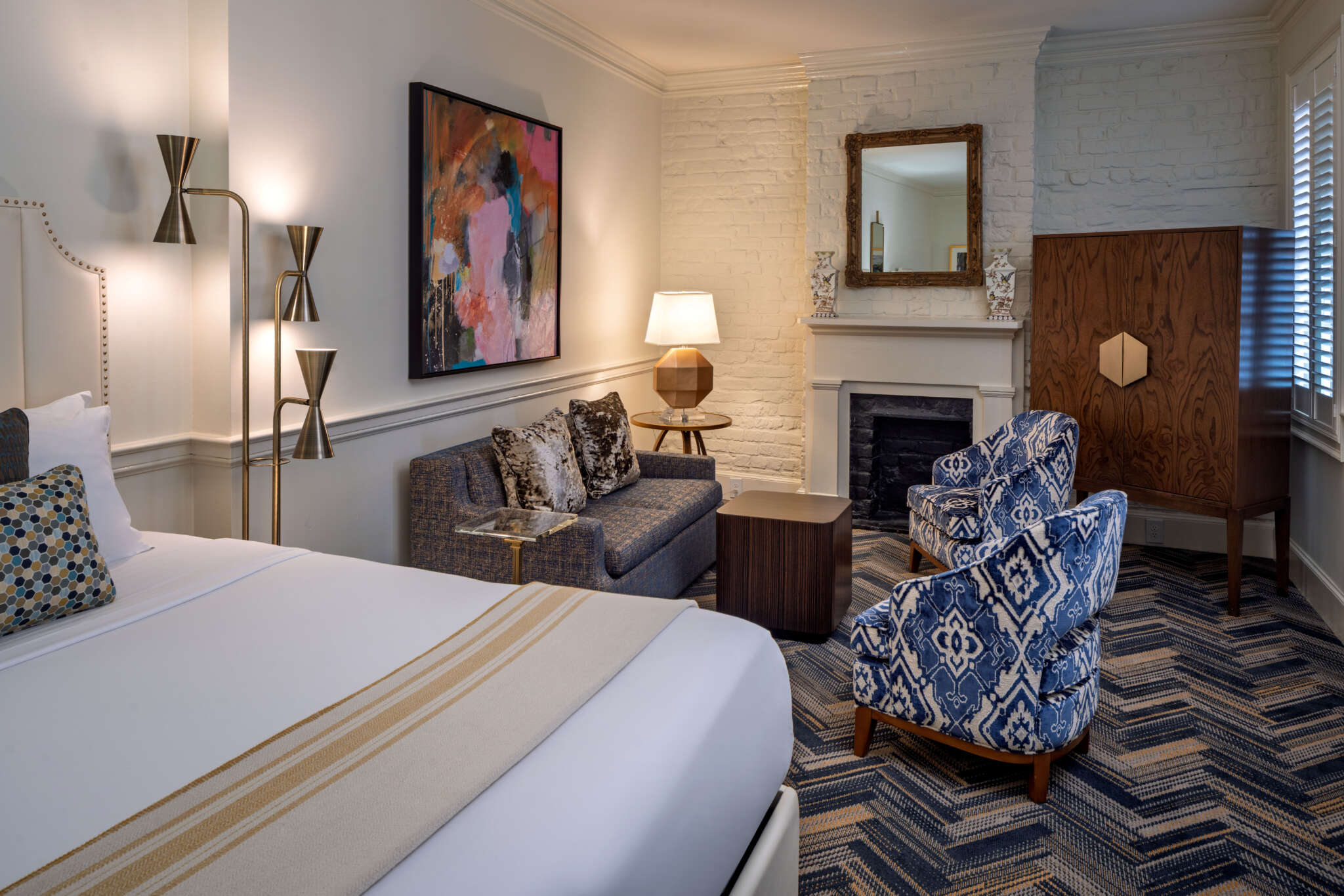 A hotel room with a bed, sofa, armchair, fireplace, and painting.